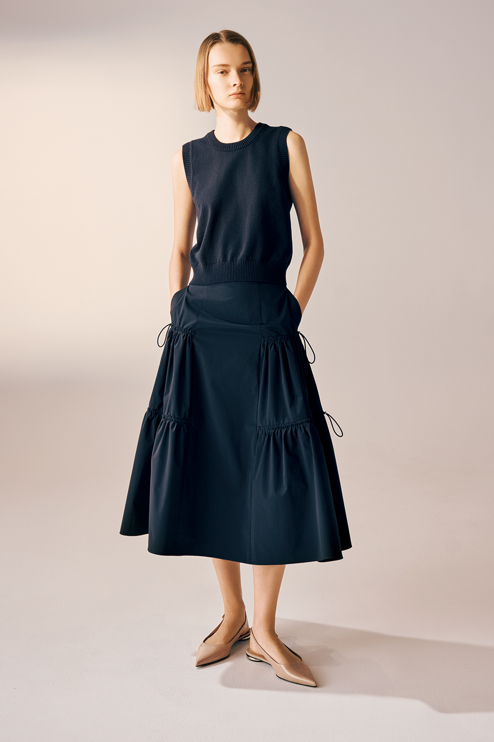 Model is wearing a navy sleeveless knit top paired with a midi-length navy skirt featuring gathered tiers and side drawstring details, styled with beige pointed flats.