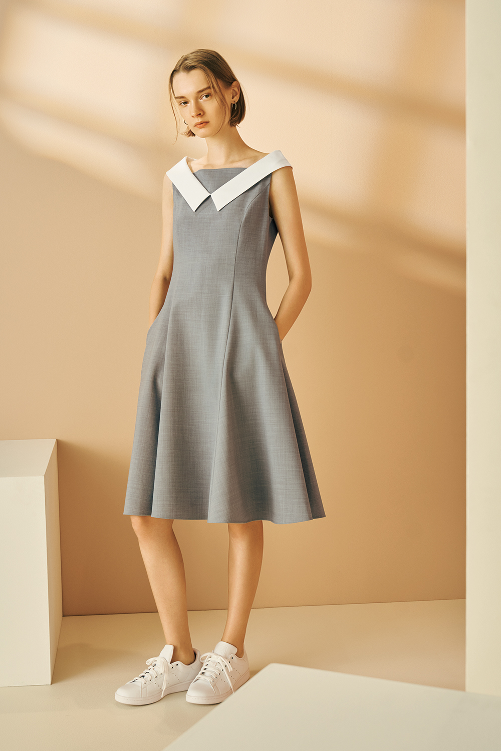 Model is wearing a sleeveless gray A-line dress with a wide white collar and white sneakers.