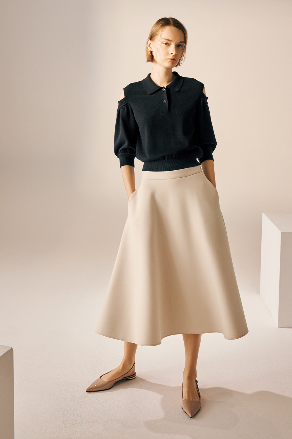 Model is wearing a dark navy short-sleeve knit polo top with shoulder cutouts, a beige A-line midi skirt, and pointed nude flats.