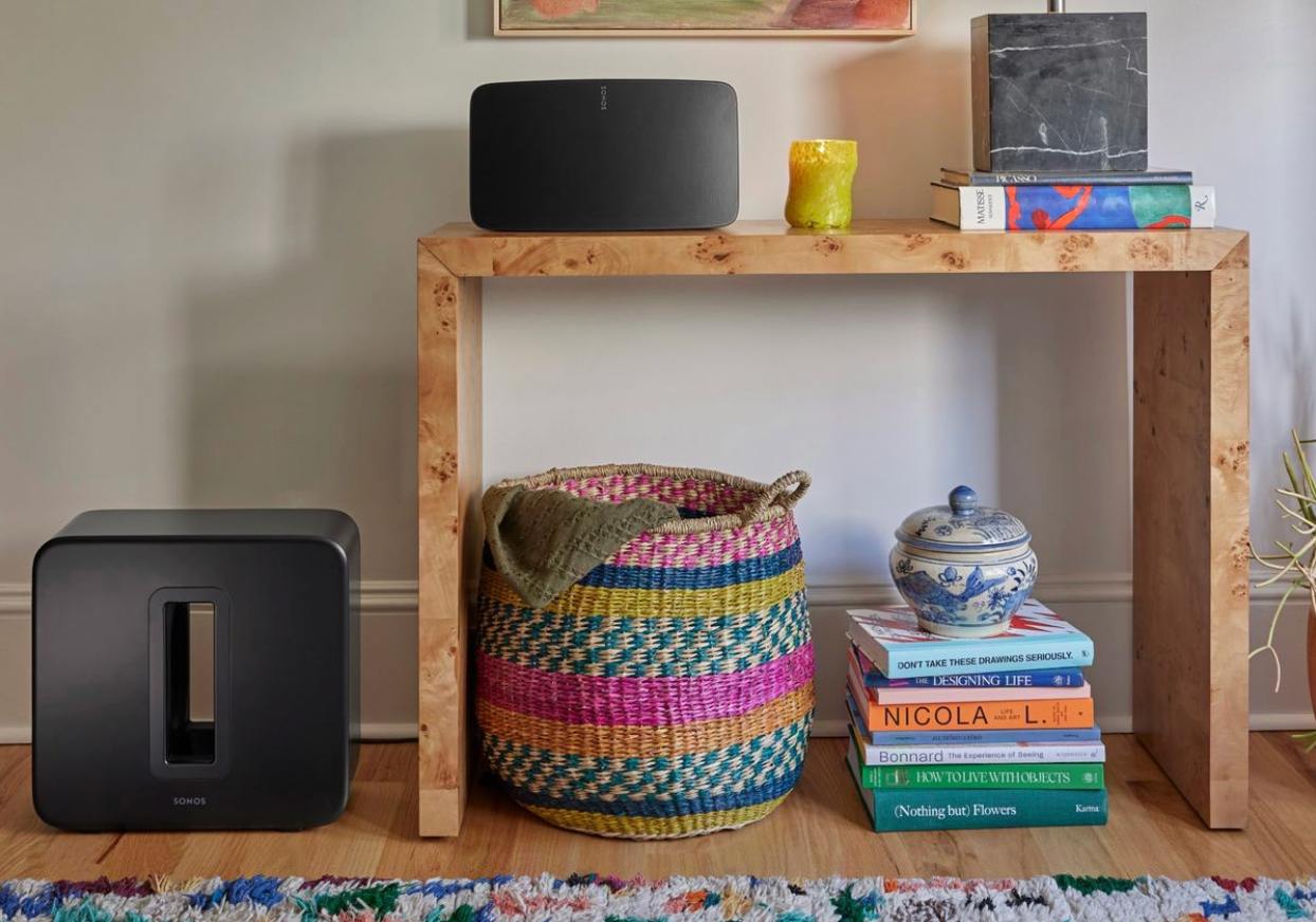 A wooden console table holds a Black Sonos Five speaker, candle, and books. Below, a colourful basket with a blanket, a Sonos Sub 4 on the left, and a ceramic jar on books to the right sit on a vibrant rug.