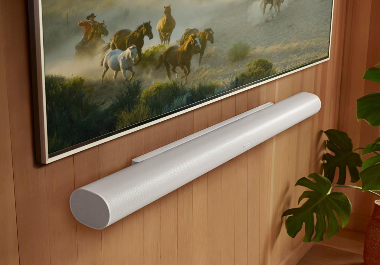 A White Sonos Arc Ultra soundbar mounted on a wall beneath a framed picture, with wood paneling and plant leaves nearby.