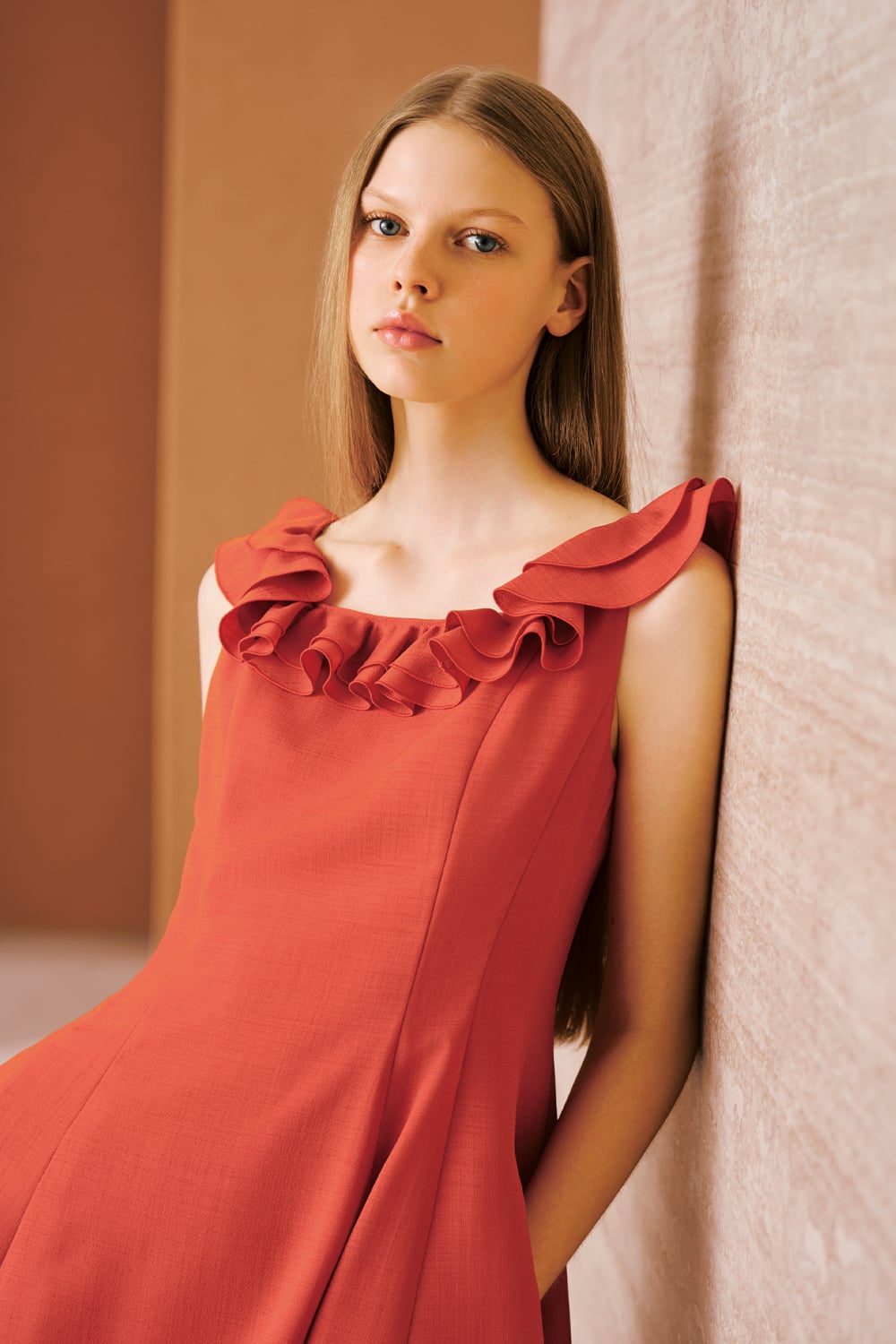 A model is wearing a coral sleeveless midi dress with ruffled detailing along the neckline.