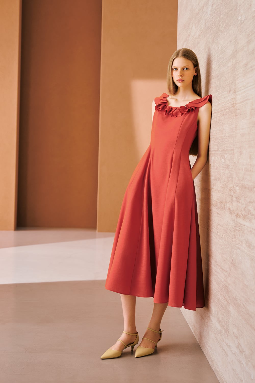 A model is wearing a coral sleeveless midi dress with ruffled detailing along the neckline and pointed heels.