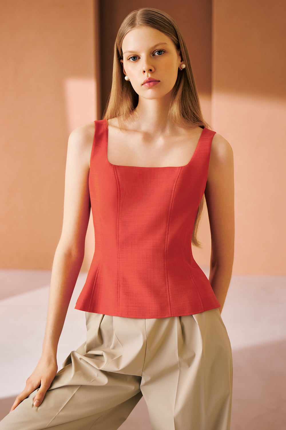 A model is wearing a coral sleeveless top with a square neckline, paired with beige wide-leg trousers.