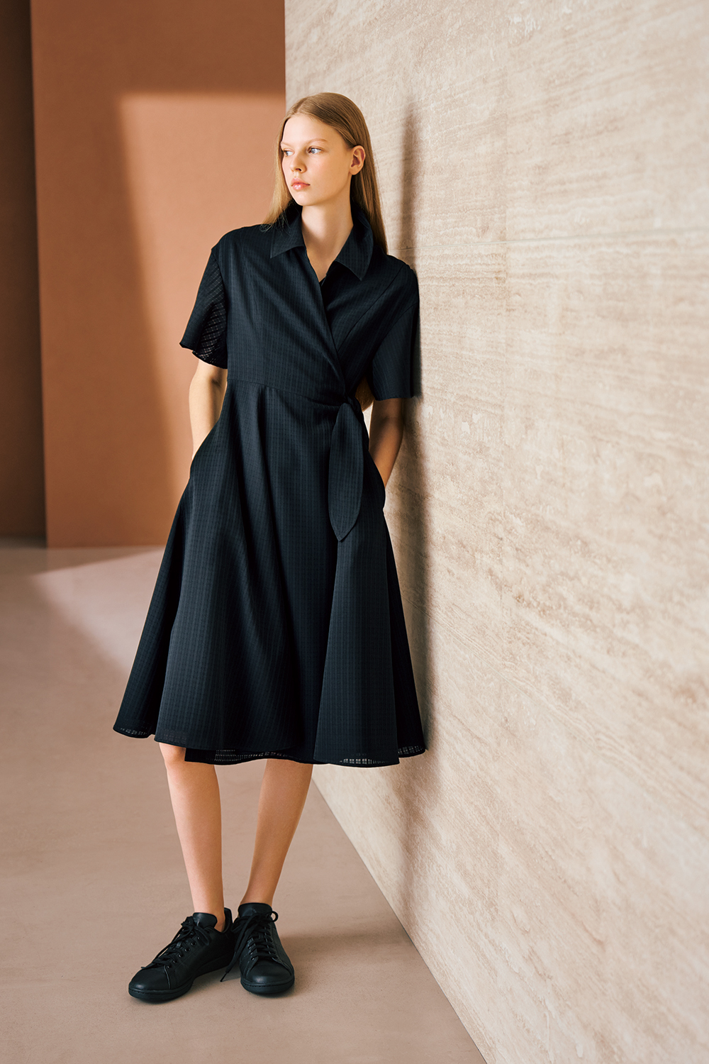 A model is wearing a black short-sleeve wrap dress with a tied waist and black sneakers.