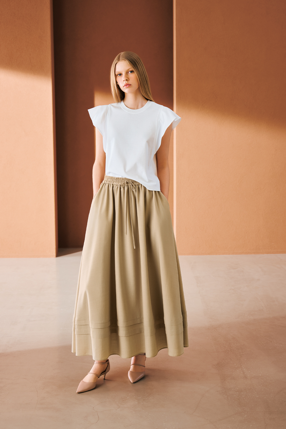A model is wearing a white sleeveless top paired with a beige gathered maxi skirt and pointed heels.