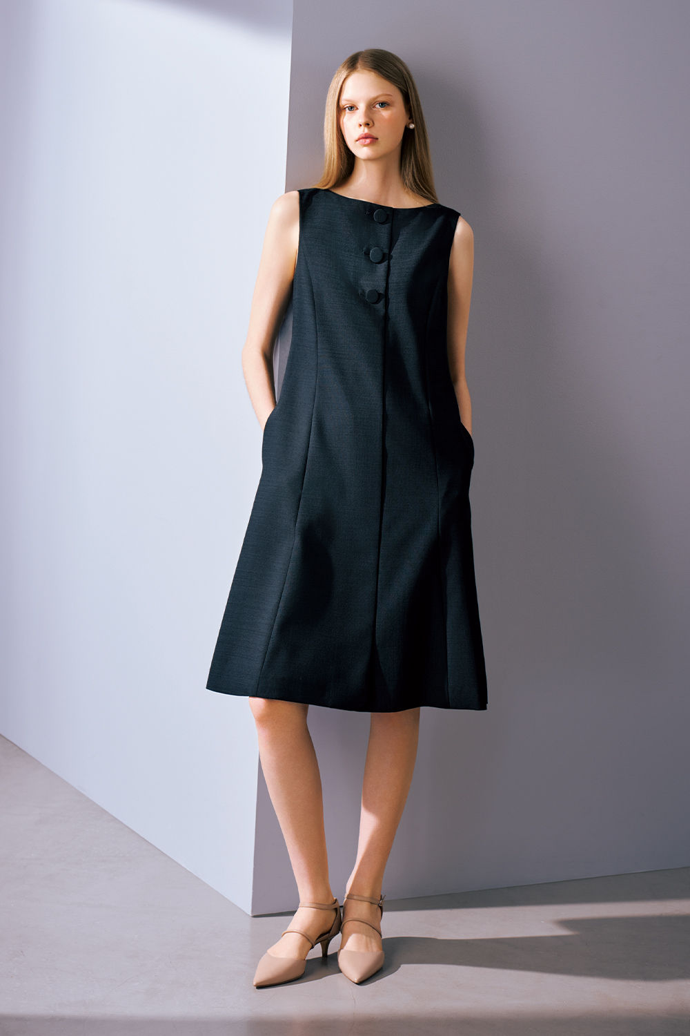 A model is wearing a black sleeveless A-line dress with oversized button detailing and pointed heels.