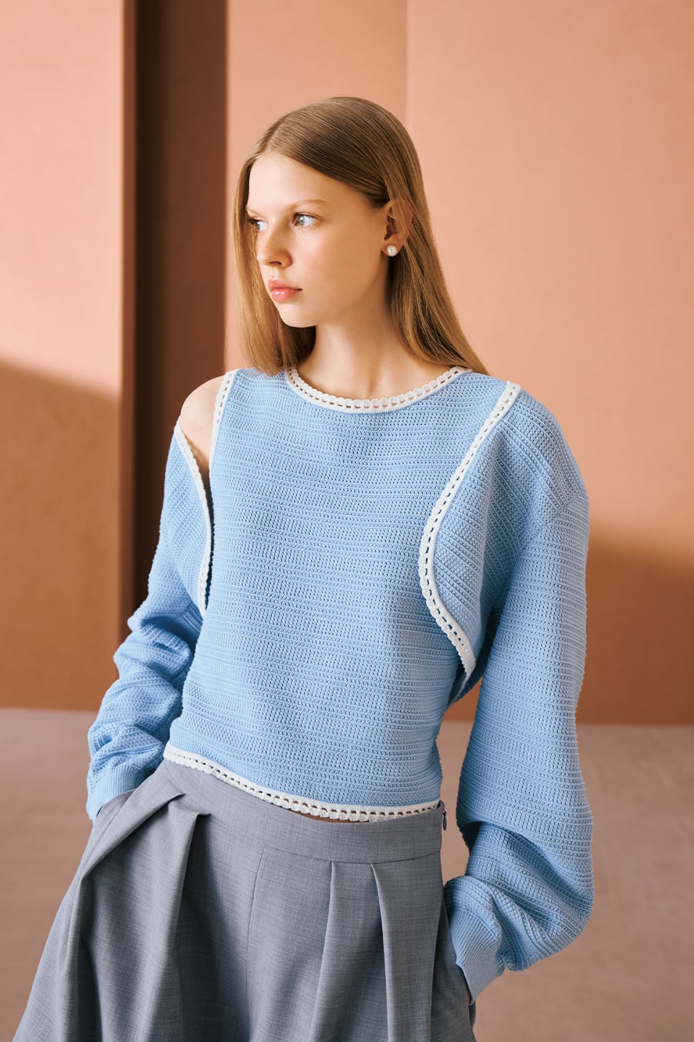 A model is wearing a light blue knit top with cutout shoulders and grey wide-leg trousers.