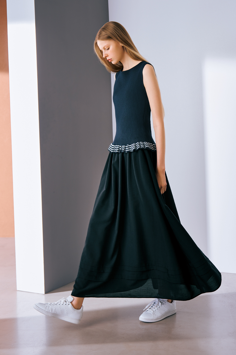 A model is wearing a navy sleeveless knit dress with striped ruffle detailing at the waist and white sneakers.