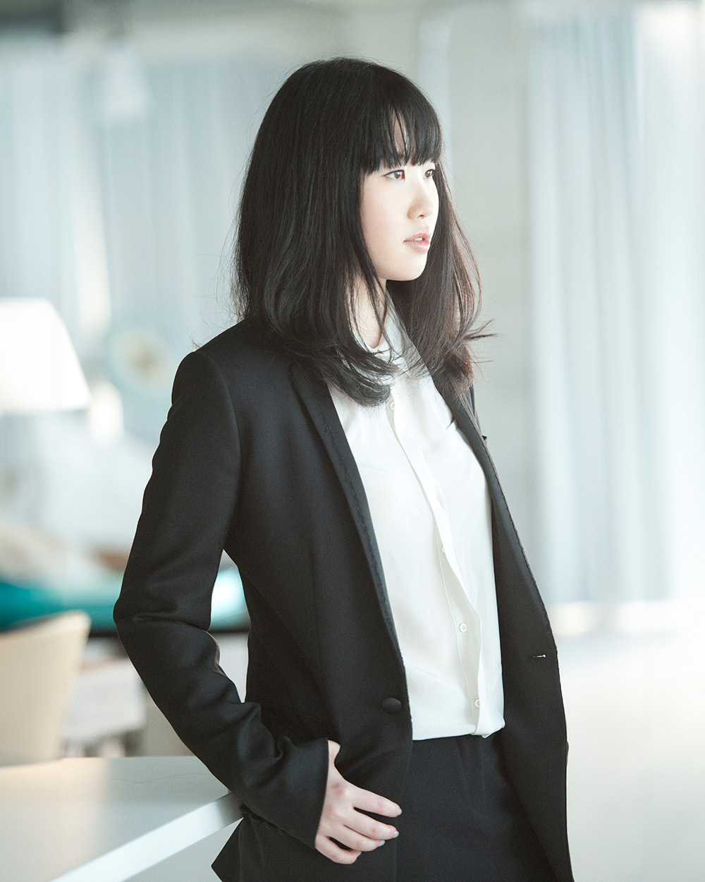 A woman in a black suit and white shirt standing indoors, looking to the side.