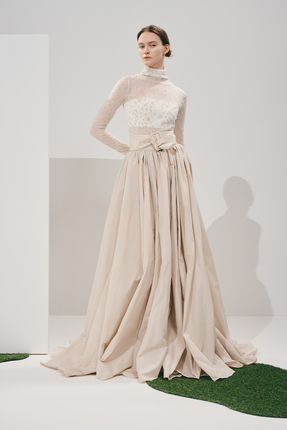 Model in a sheer high-neck floral blouse and voluminous cream pleated maxi skirt with a wide matching belt, in a white studio with patches of green turf.