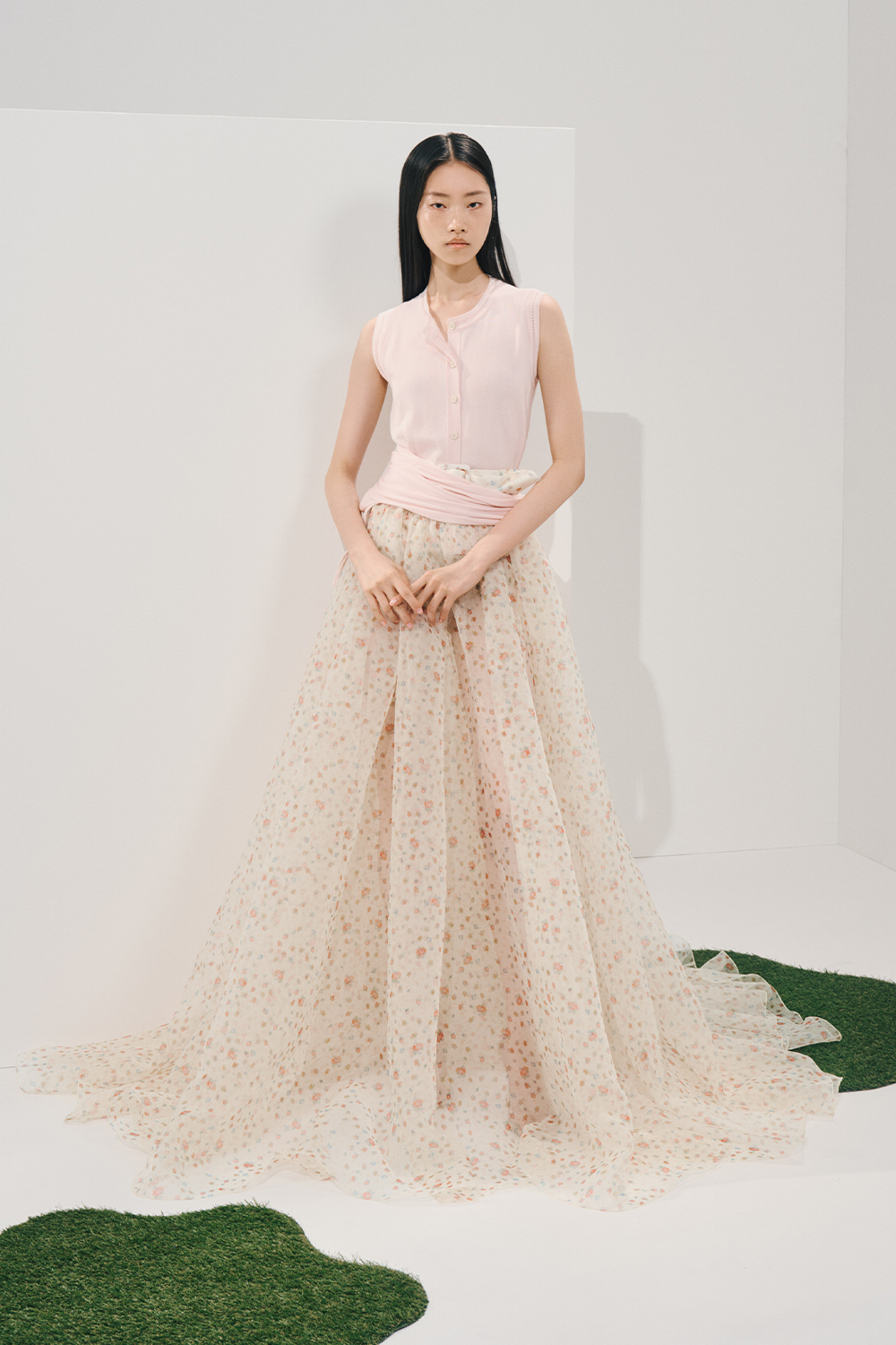 Model in a sleeveless light-pink ribbed knit top with a matching sash and a sheer cream floral maxi skirt, standing in a minimalist white studio.