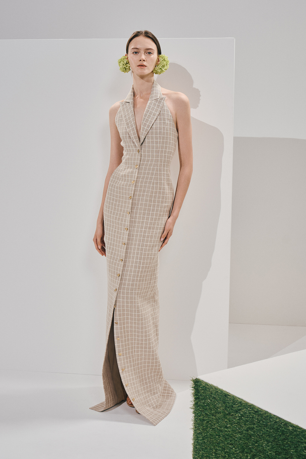 Model in a sleeveless beige windowpane maxi dress with a notched collar and gold buttons, wearing large light-green floral pom-poms over the ears.