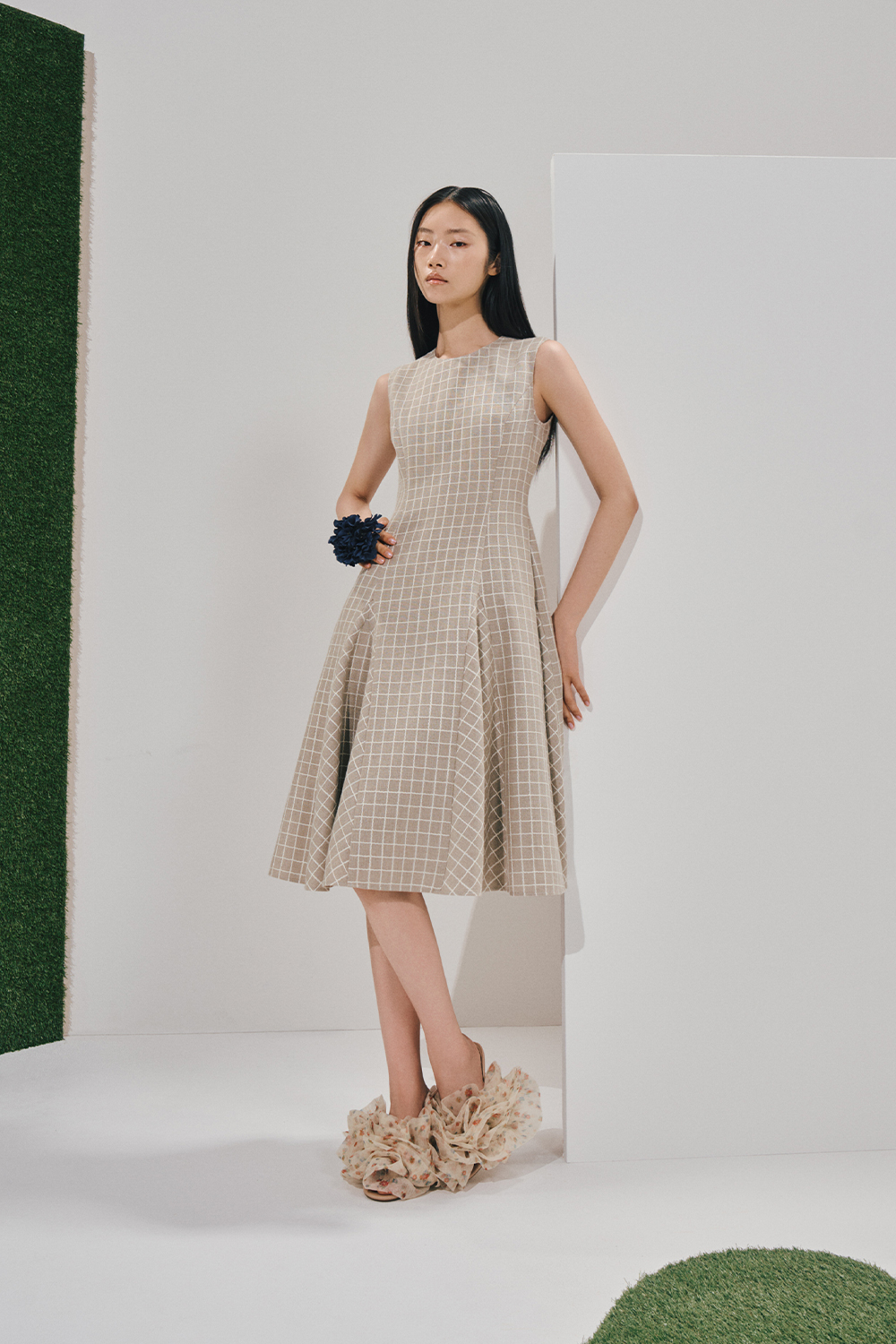 Model in a sleeveless beige windowpane A-line dress, a navy ruffled wrist accessory, and cream floral ruffled slide sandals, leaning on a white studio panel.