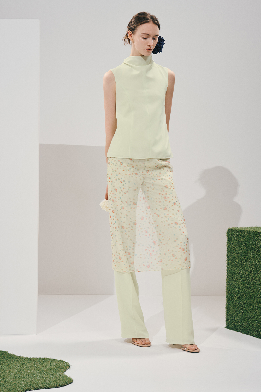 Model in a pale mint mock-neck top and matching wide-leg trousers under a sheer floral midi overskirt, with a navy floral hair accessory and nude sandals.