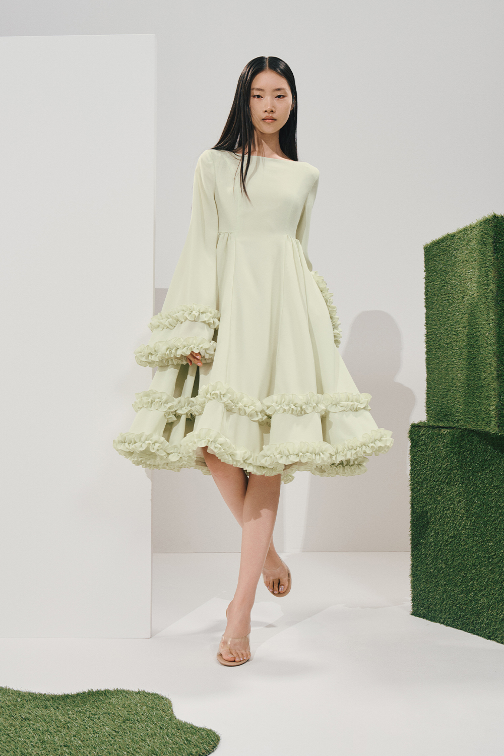 Model in a pale mint fit-and-flare dress with voluminous bell sleeves and tiered ruffles at the hem, worn with clear heeled sandals beside stacked turf cubes.