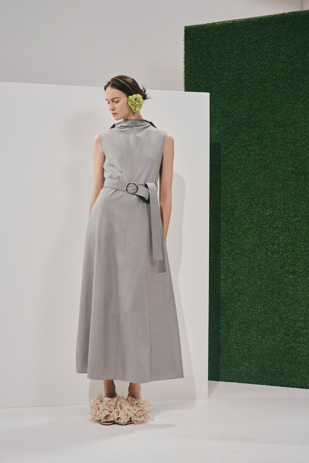 Model in a sleeveless light-grey mock-neck maxi dress with a matching belted waist, ruffled cream slides, and a large light-green flower behind the ear.