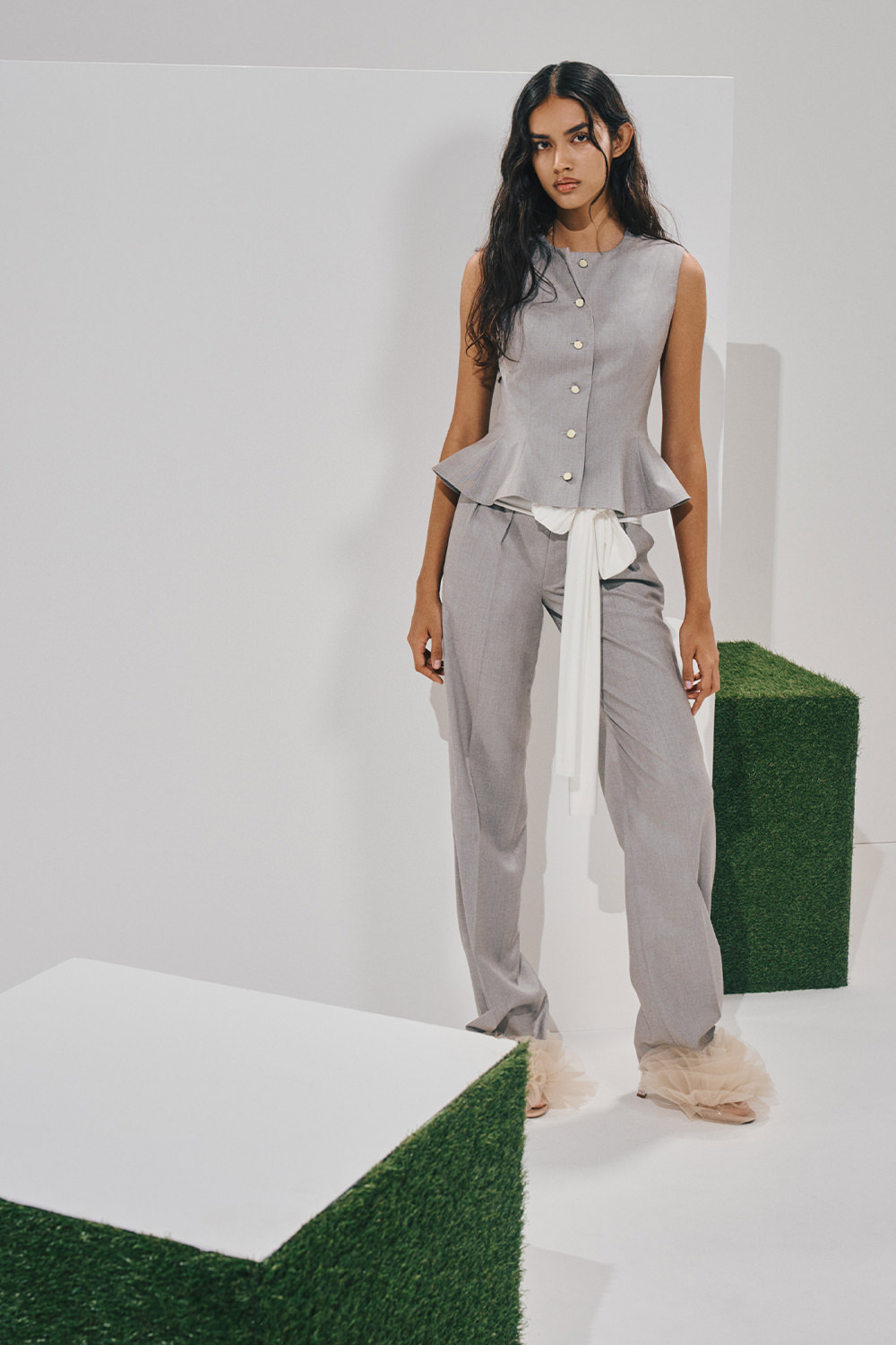 Model in a light-grey sleeveless peplum vest and matching straight-leg trousers, tied with a long white sash, and ruffled high-heeled sandals.