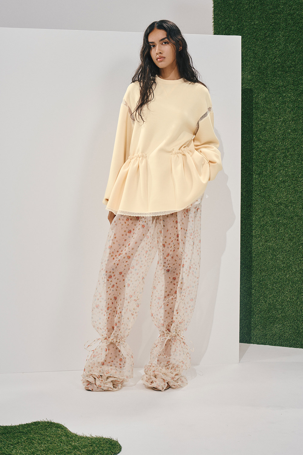 Model in an oversized cream sweatshirt with lace-trimmed peplum hem and sheer floral wide-leg trousers gathered at the ankles with ribbons.