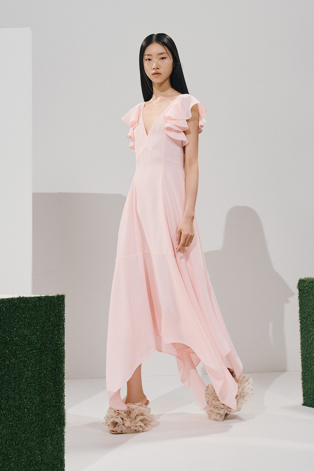 Model in a pale pink ruffled midi dress with a V-neck, flutter sleeves, and an asymmetrical handkerchief hem, worn with voluminous cream floral ruffled sandals.