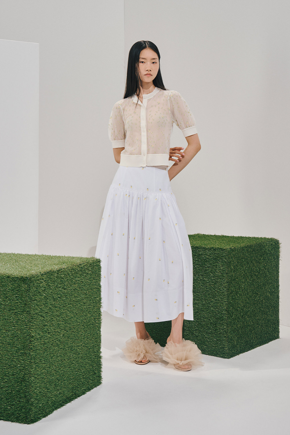 Model in a sheer cream floral short-sleeve cardigan over a white midi skirt with sparse yellow floral embroidery, and beige voluminous ruffled slide sandals.