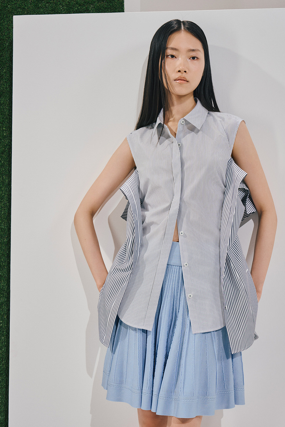 Model in a sleeveless light-blue pinstripe shirt with structured wing-like side panels and a matching accordion-pleated knee-length skirt.