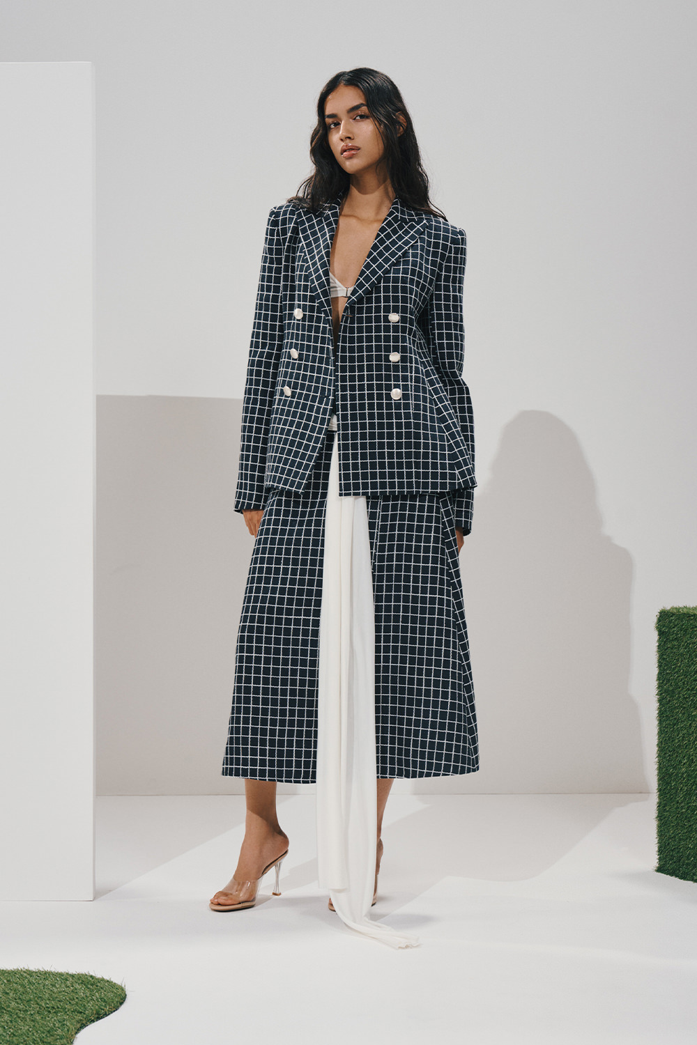 Model in a navy windowpane double-breasted blazer and matching midi skirt, with a long white draped sash and clear pointed-toe heeled mules.