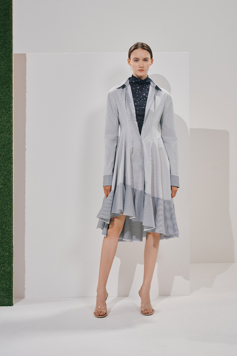 Model in a light-blue shirt dress with an asymmetrical ruffled hem layered over a navy patterned turtleneck, with clear heeled sandals.