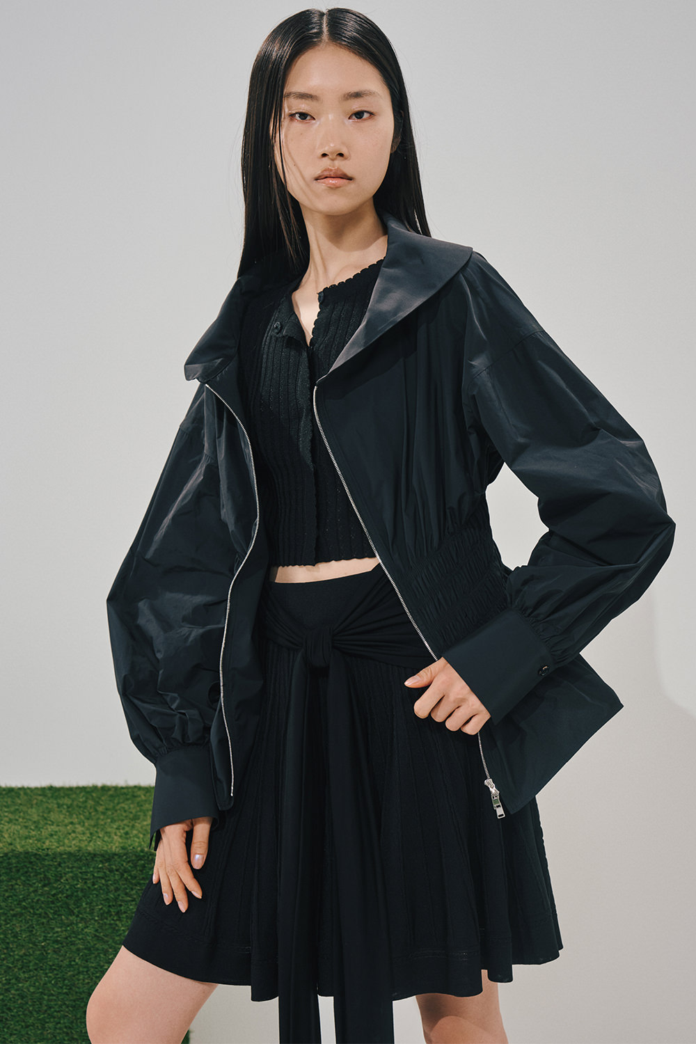 Model in an all-black look: an open zip jacket with voluminous puff sleeves, a ribbed cropped button top, and a pleated skirt with a fabric tie belt.