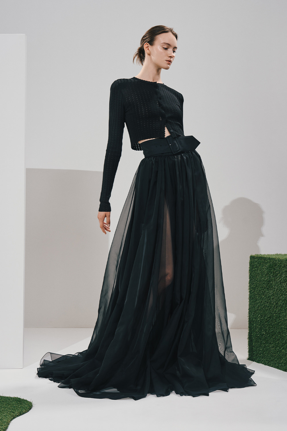 Model in a black cropped ribbed knit top and a voluminous sheer black maxi skirt cinched with a wide belted waist, in a white studio with green turf props.