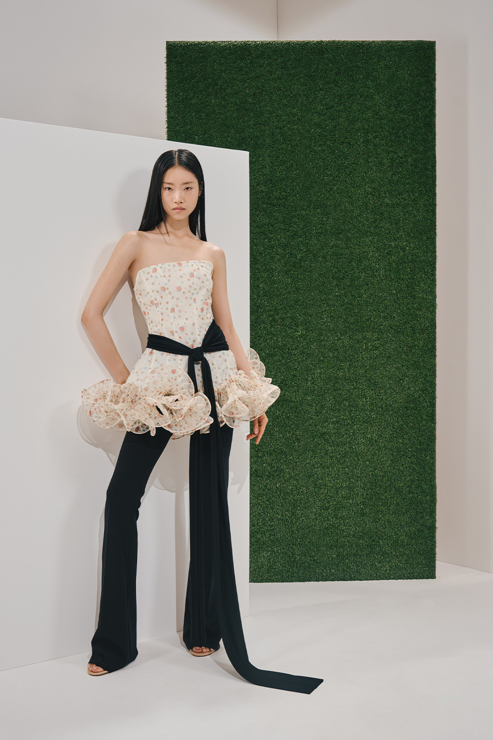 Model in a cream floral strapless bodice with large hip ruffles, a long black sash trailing to the floor, and slim black trousers.