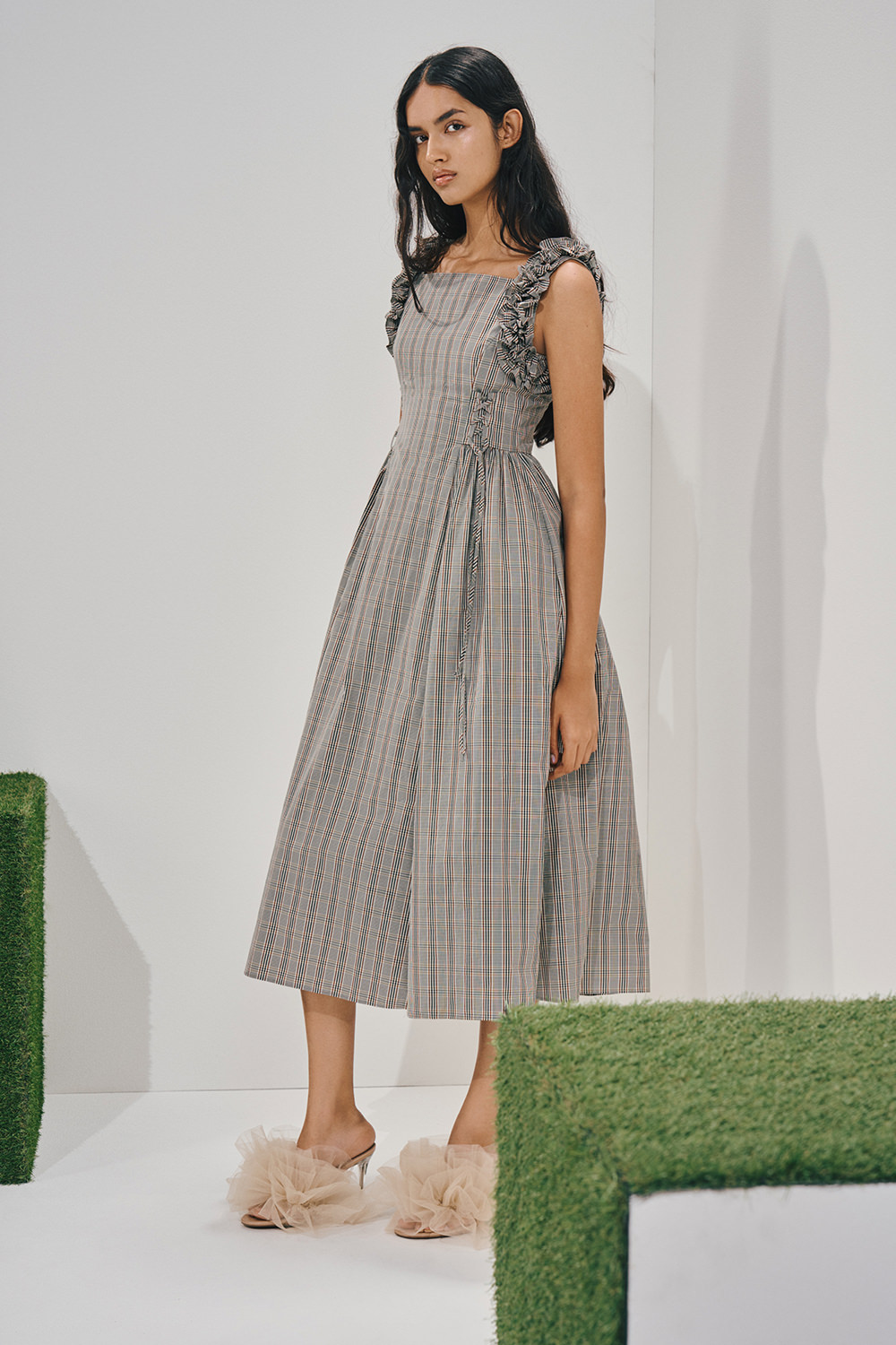 Model in a sleeveless grey-blue plaid midi dress with ruffled straps, a side corset lace-up, and an A-line pleated skirt, worn with voluminous ruffled sandals.
