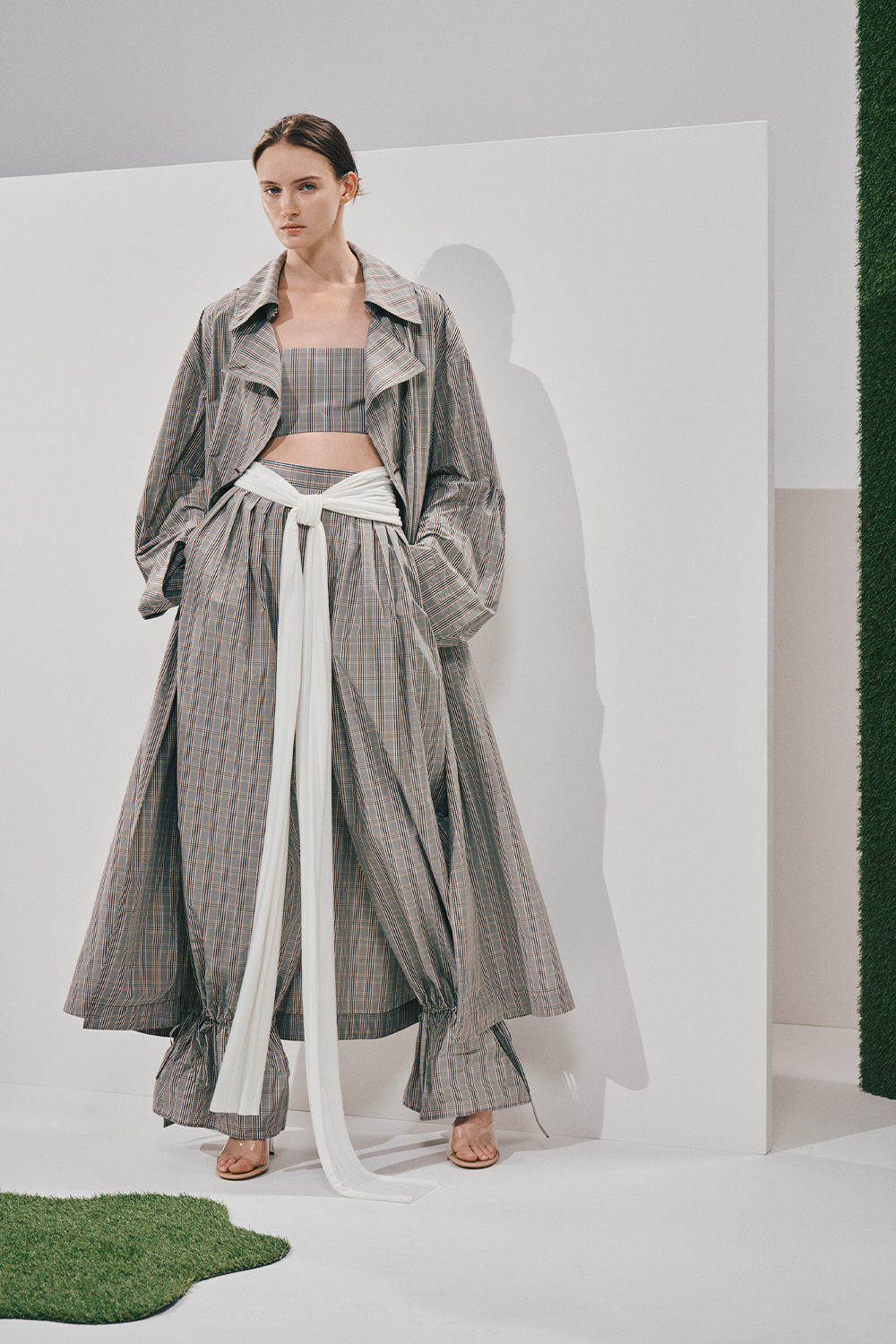 Model in a matching grey plaid set: an oversized trench coat, strapless crop top, and wide-leg trousers gathered at the ankles, tied with a long white sash.