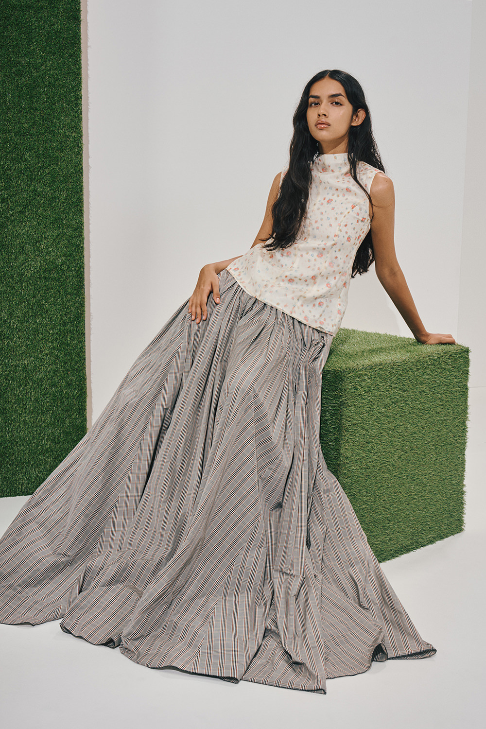 Model in a white sleeveless floral high-neck top and a voluminous grey plaid floor-length skirt, leaning against a green turf cube in a white studio.