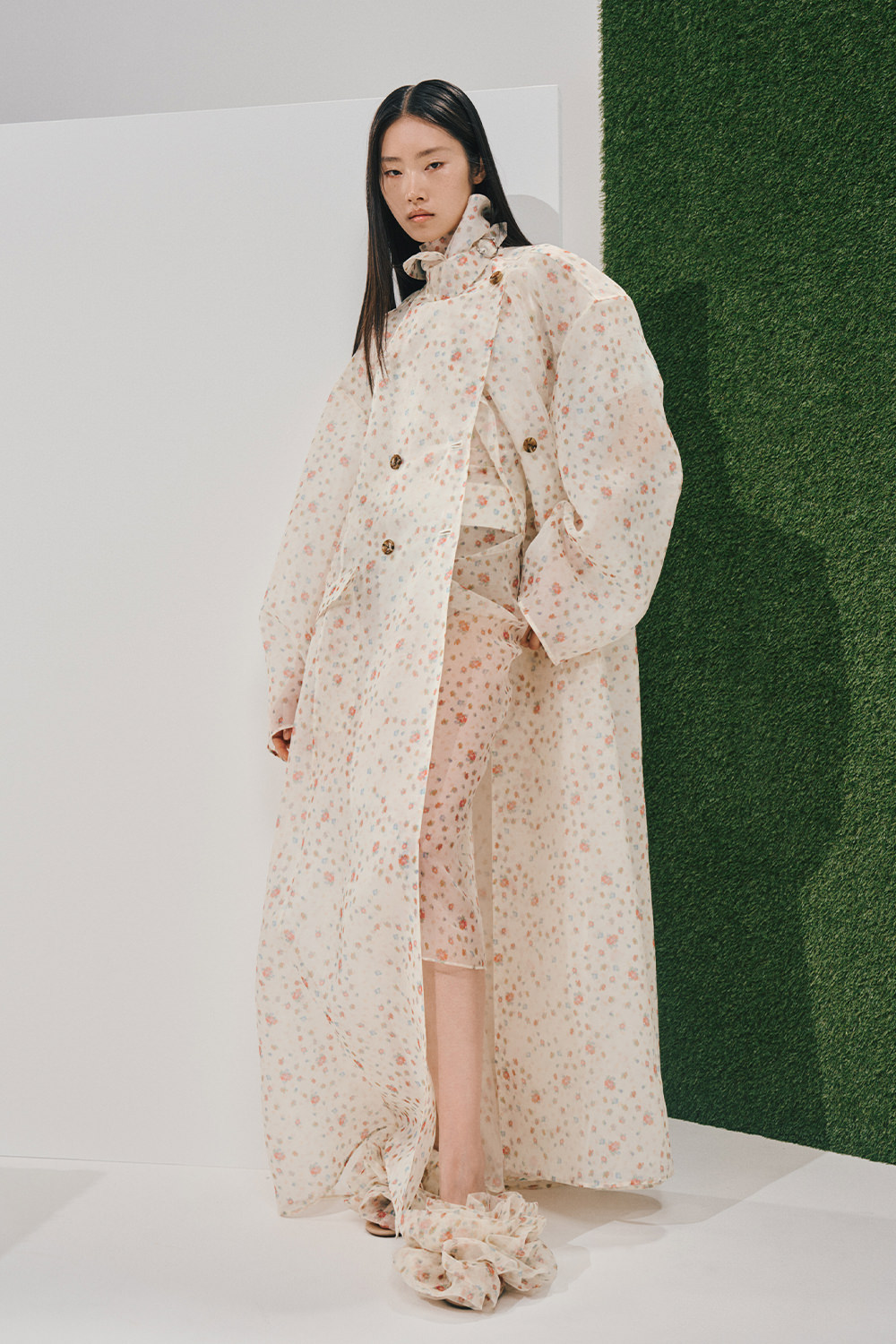 Model in a sheer off-white floral floor-length double-breasted coat with balloon sleeves over a matching floral top and skirt, with coordinating ruffled sandals.