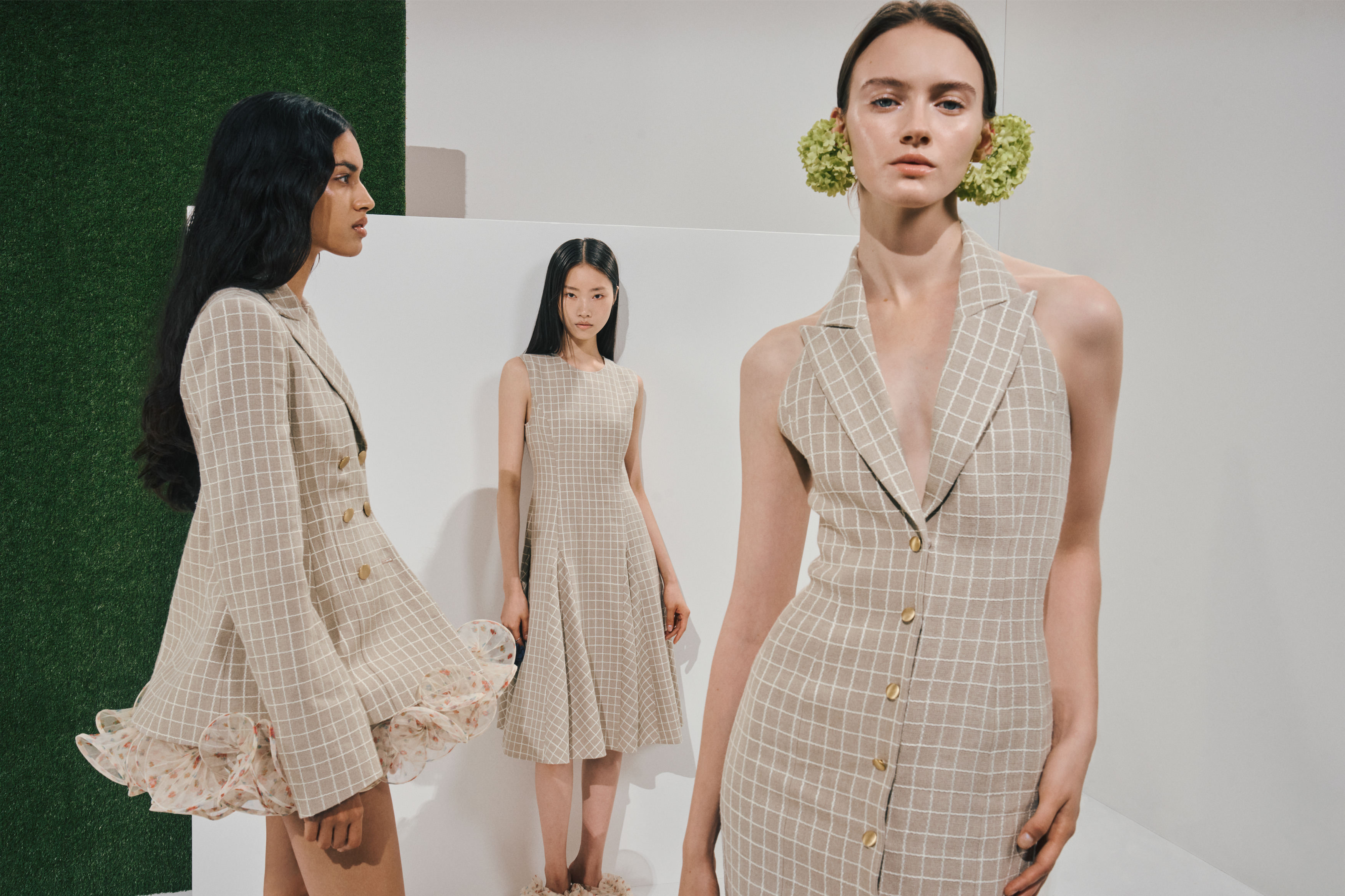 Three models in matching beige windowpane looks: a sleeveless halter-neck vest dress with gold buttons and green hydrangea earrings, a double-breasted blazer with a floral ruffled hem, and a sleeveless A-line dress, in a white studio with a green turf panel.