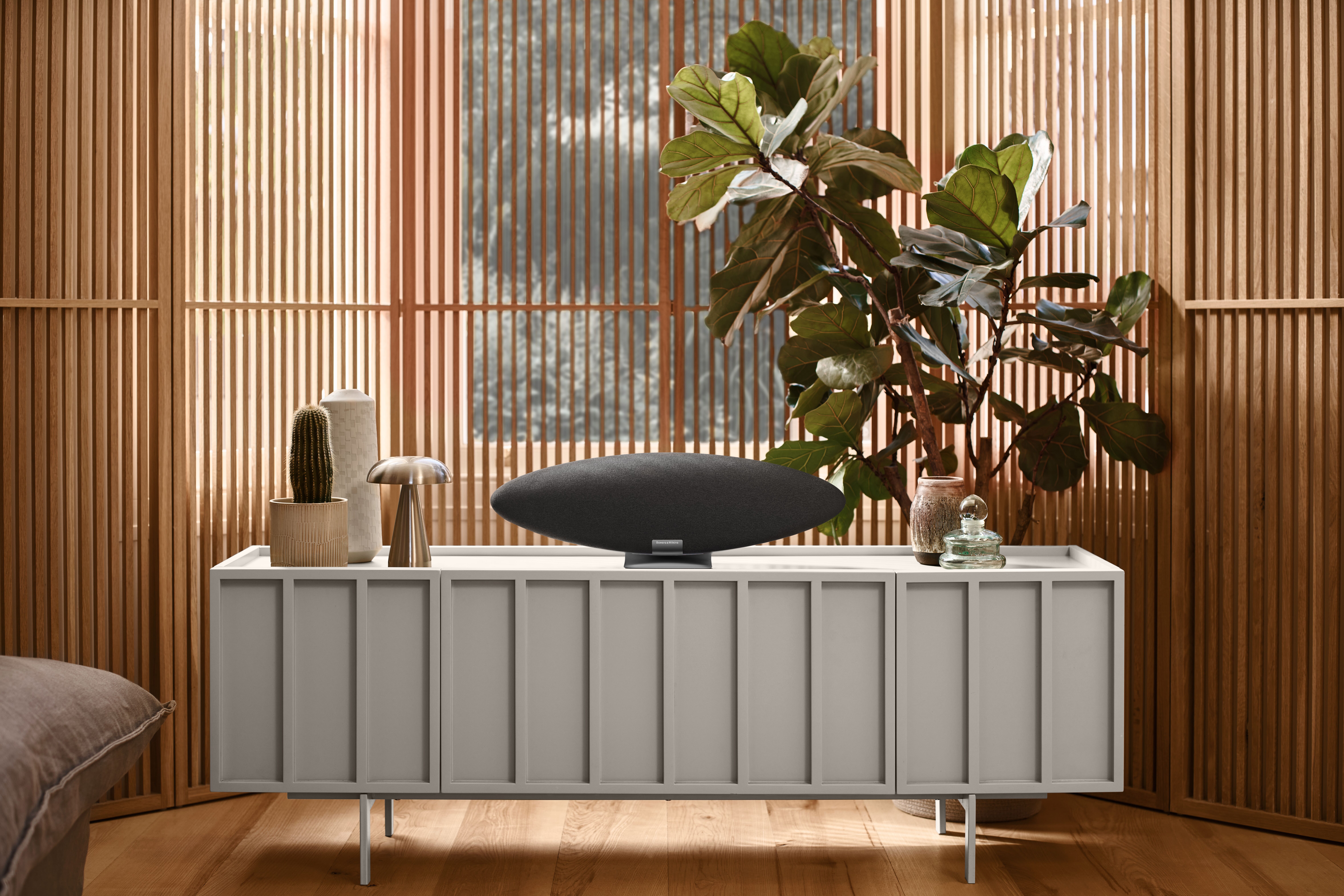Bowers & Wilkins Zeppelin Pro Edition wireless speaker in Space Grey displayed on a modern console table in a contemporary living room with wood paneling and indoor plants.