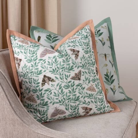 Two pale pink and green scatter cushions with moth, insect and floral designs, arranged on a light grey armchair in front of a neutral background.
