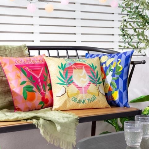 Three colourful outdoor cushions with cocktail themed designs, propped up on a wooden bench in an outdoor setting.