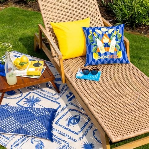 A garden seating area decorated with a woven sun lounger, bright blue and yellow garden cushions and a tropical outdoor rug.