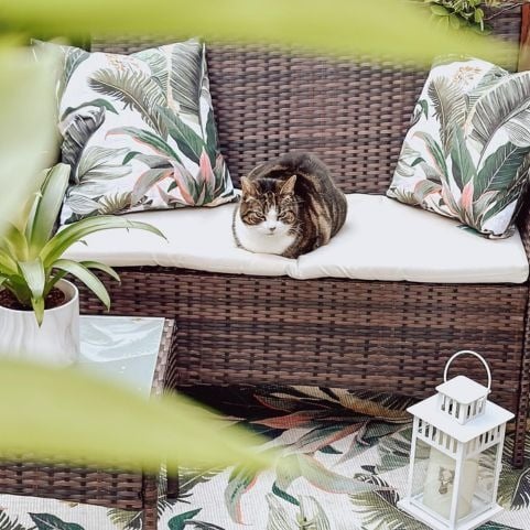 A garden seating area decorated with a woven outdoor sofa, outdoor cushions with a leafy tropical design and a matching garden rug.