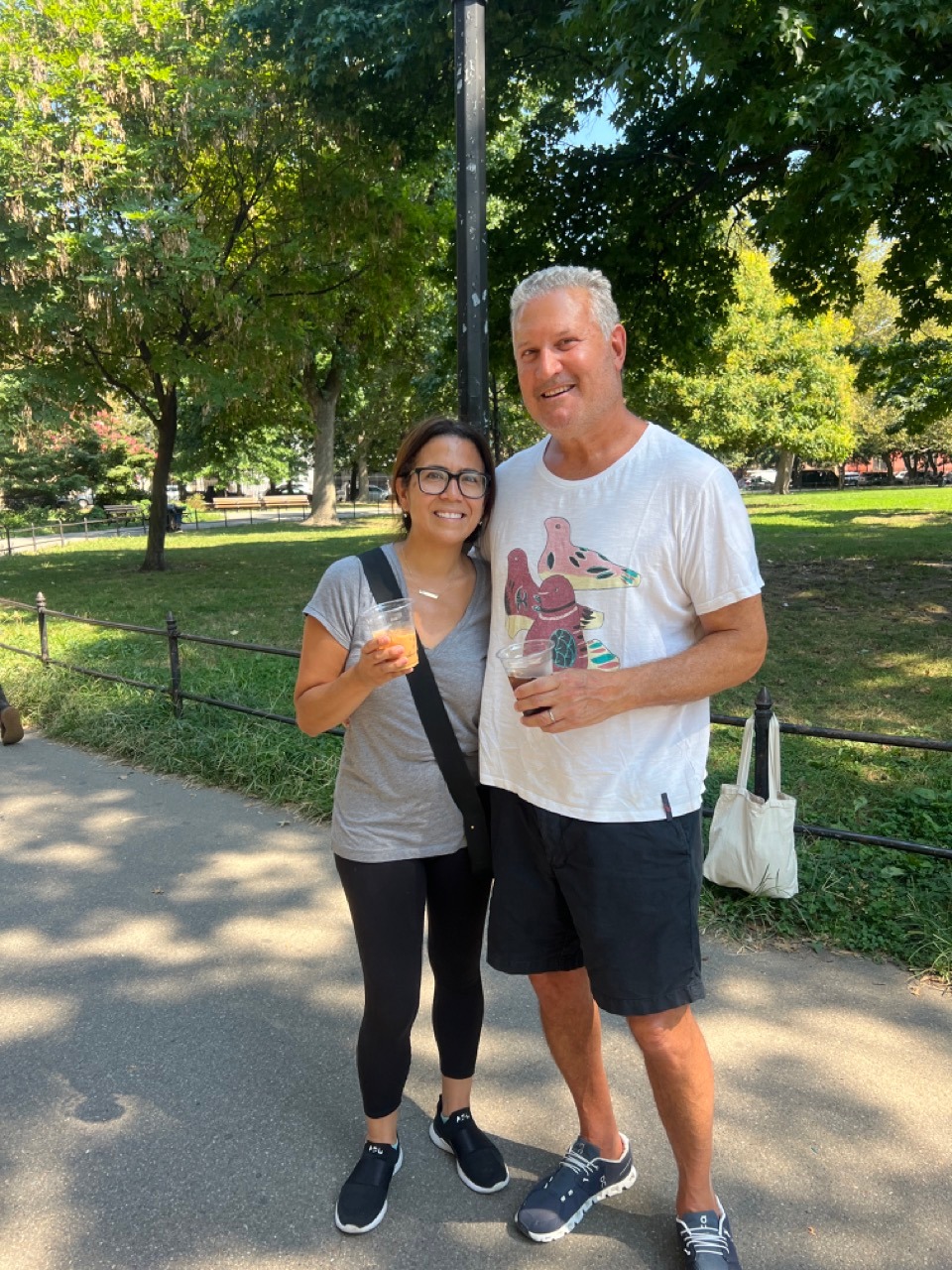 Two people stand in the park smiling with their coffees.