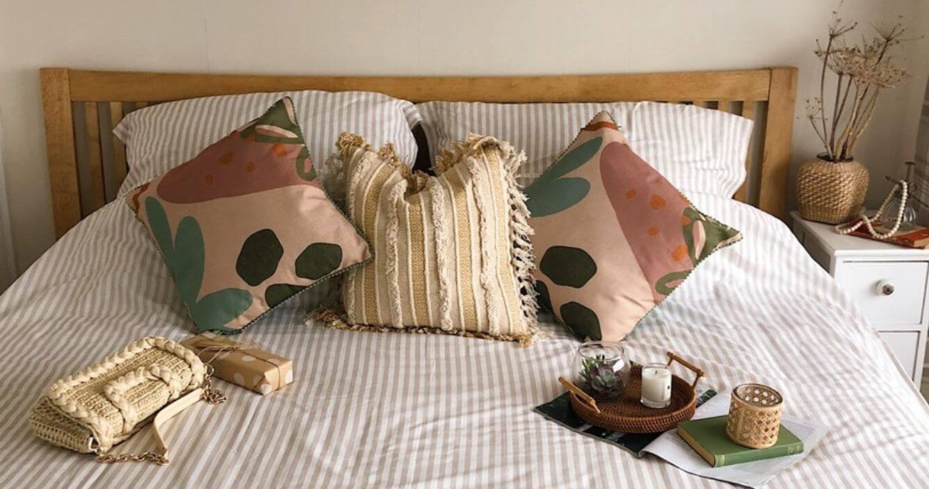 A bed piled high with cushions in a relaxed style. The bedding is pale, and the cushions have earthy tones. On one side of the bed there is a tray with a candle and a terrarium, and on the other a rattan handbag and a wrapped present. 