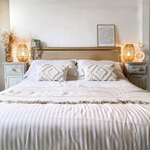 A light, scandi feel bed with a light wood headboard. The bedding is off white stripe, the two cushions in front of the pillows are cream and tufted in a diamond pattern, and the throw is textured and fringed. 