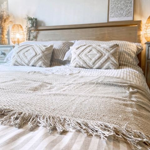A light, scandi-feel bed. We have a close up on the throw. It is cream-coloured, textured and fringed along the edge. In the background, there is two rectangular tufted cushions, a striped bedding and a light wood headboard.