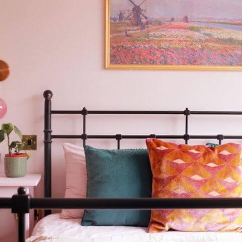 A black metal headboard dressed with a selection of beautiful cushions. The wall behind the headboard is pink, with a painting of a dutch flower field scene hung above the bed. On the bed there is a dusky teal cushion and an orange and pink velvet cushion.