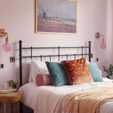 A pink bedroom with a black metal bed making the focal point of the image. The bed is dressed in a selection of beautiful cushions and throws. There is a cylindrical bolster cushion in pink, two dusky teal square cushions and one orange and pink geometric cushion at the front and centre of the bed.