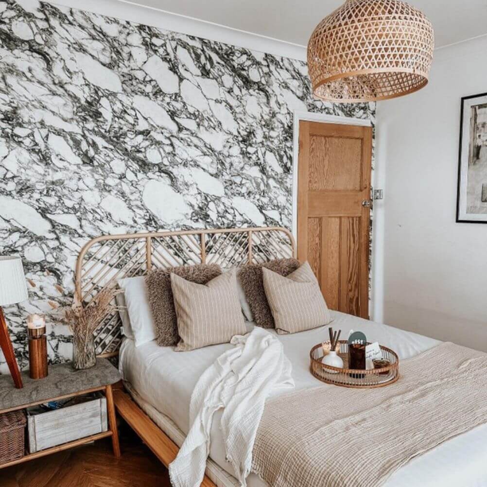 A scandi style bedroom with a striking black and white marble wallpaper feature wall. The headboard is rattan, as is the light fixture. The bed is dressed in tonal beige and latte hues, with a set of two faux fur cushions. On the bed there is a rattan tray with a candle, card and more on. 