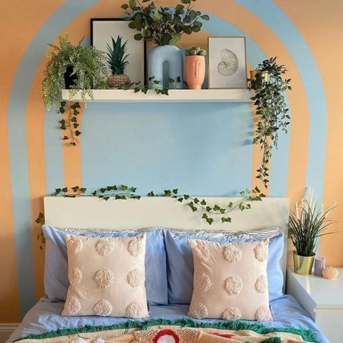 A summery bedroom with a painted on arch in blue acting as a focal point for the wall behind the bed. There is a shelf above the bed filled with plants and artwork. The bed is dressed in blue bedding with tufted cushions.