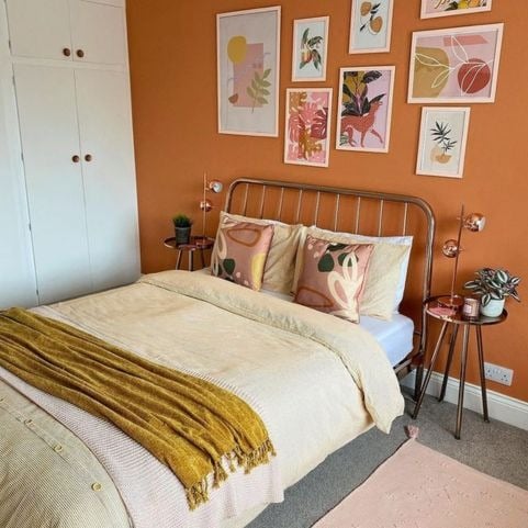 An orange bedroom with a gallery wall above the bed, featuring illustrative artwork of leaves, animals, fruits and more. The bed has a brass metal frame, light yellow bedding, abstract leafy patterned cushions, and a mustard yellow throw.